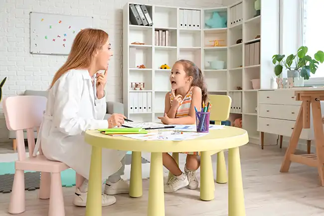 Kvinnelig logoped viser en liten hvordan forme en lyd. De sitter ved et lite gult barnebord med bokhylle med bøker i bakgrunnen